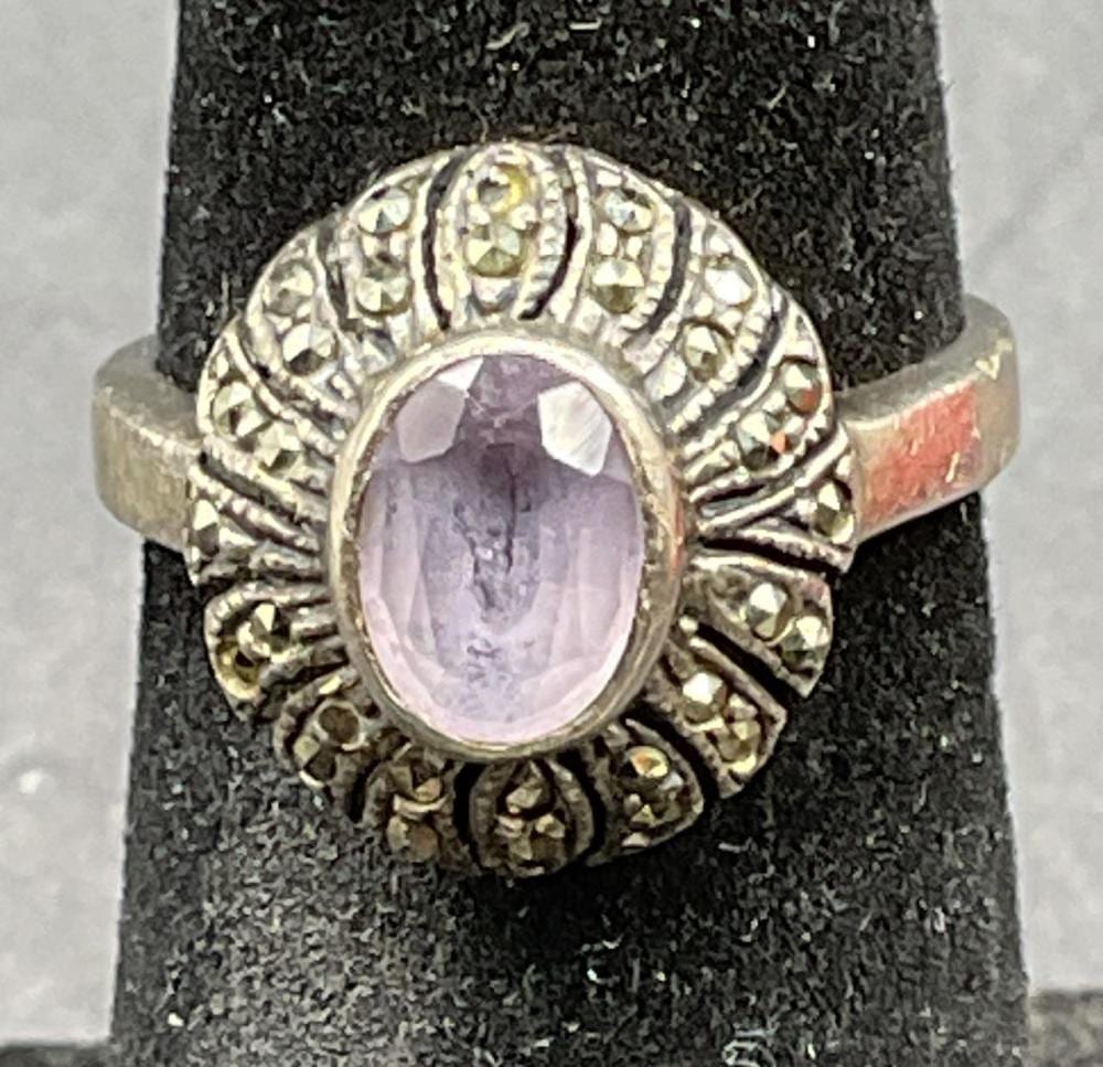 Close-up of a silver ring with an oval purple gemstone set in an ornate bezel and small patterned accents, on a dark display cushion.