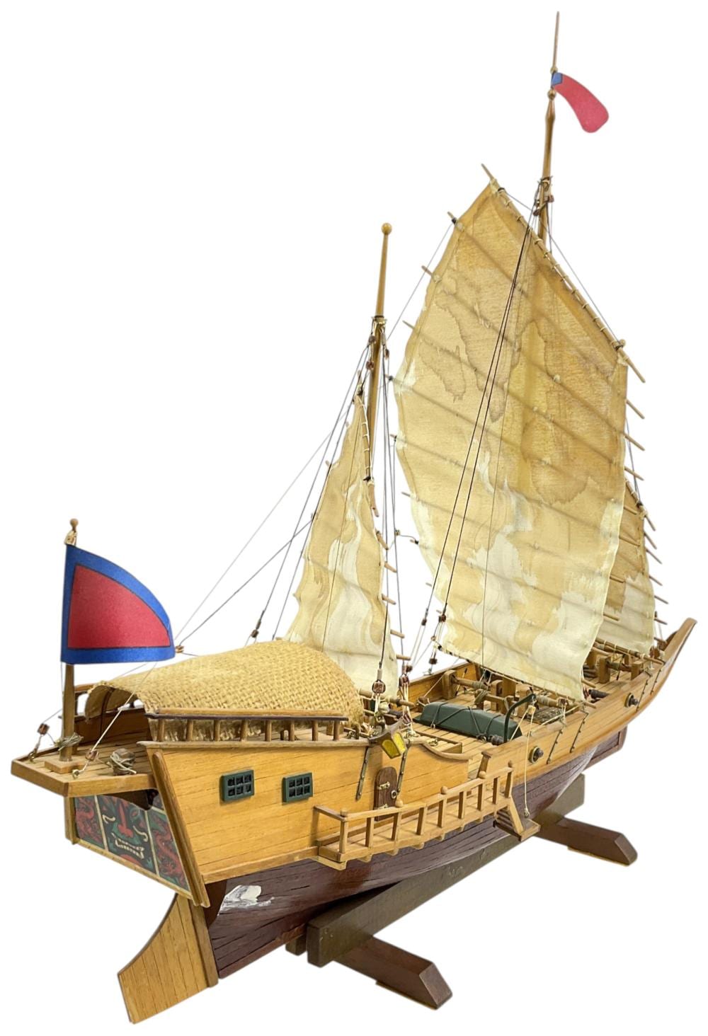 Wooden model ship on a stand with tan sails and ropes, featuring a red-and-blue flag on the left.