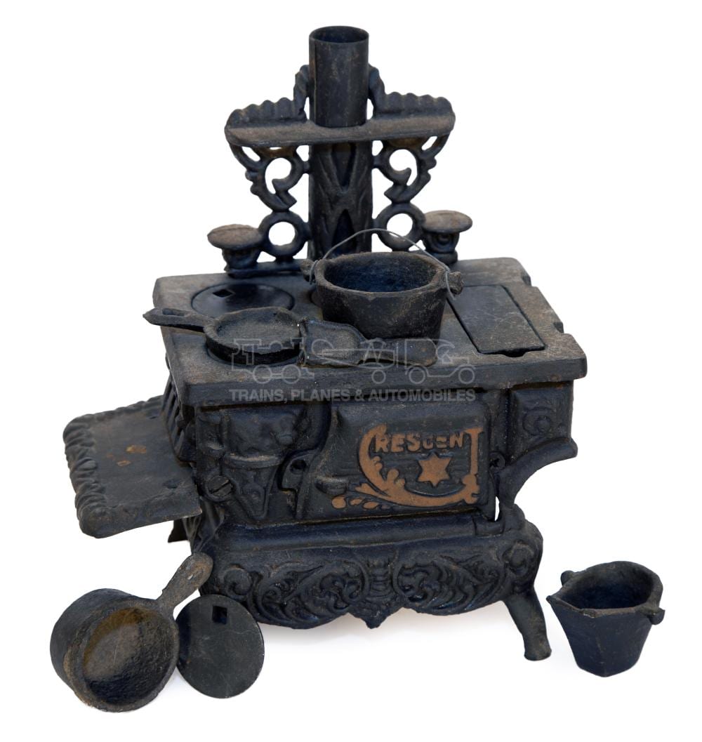 Cast iron miniature toy stove with a chimney stack, small side openings, and a front panel showing a crest, with miniature pots and tools nearby.