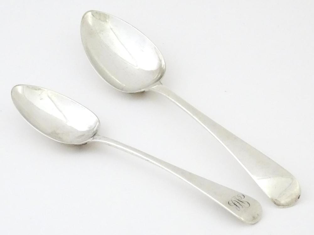 Two polished silver spoons lying on a light background, with oval bowls and short engraved handles.