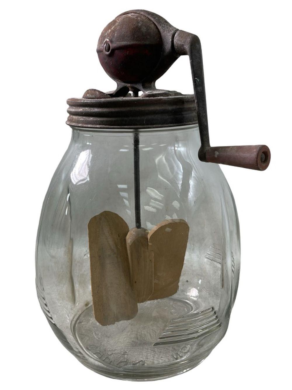 Antique glass butter churn with dark metal top and hand crank, shown upright against a white background.