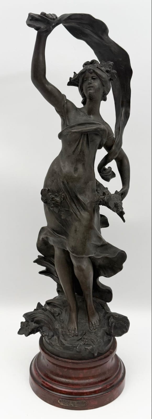 Bronze-toned statue of a nude woman holding a draped cloth above her head, standing on an ornate base.
