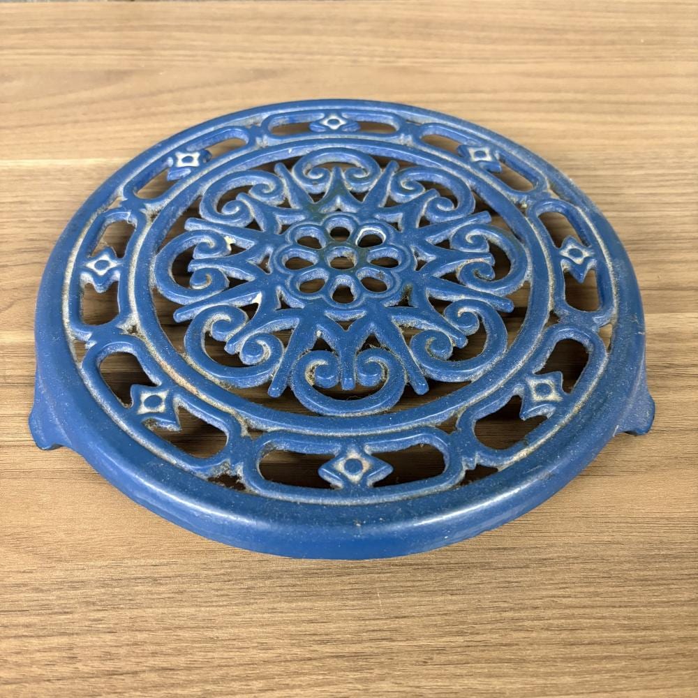 Round blue cast-iron trivet with ornate scrollwork and a central floral-style cutout pattern on a wood table.
