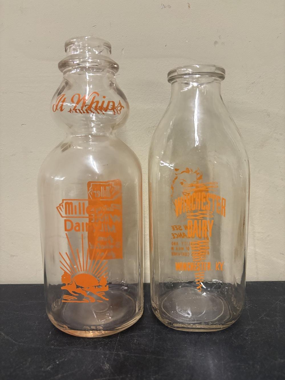 Two vintage clear glass milk bottles with orange printed labels, shown side by side against a plain wall.