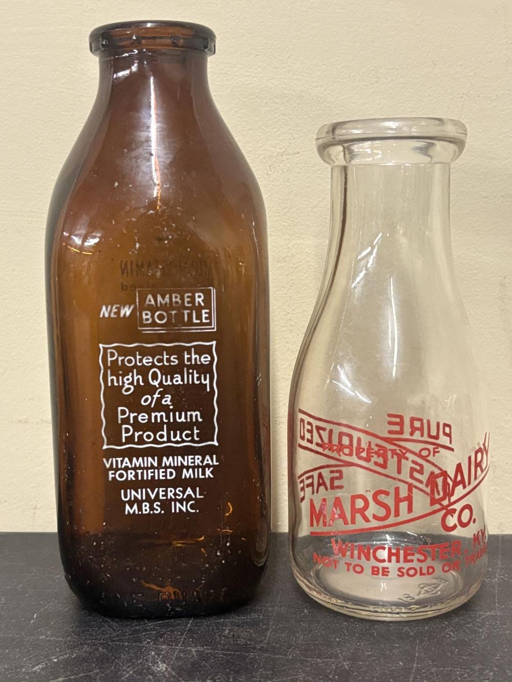 Two vintage glass milk bottles, one amber-brown with printed labels and one clear with a red label, standing side by side.