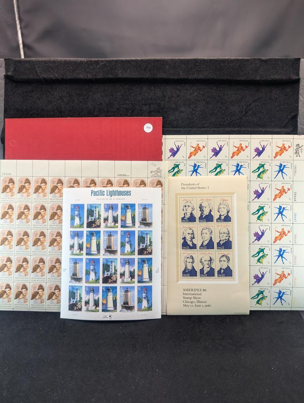 Pages of US postage stamps in rows, with a red cover page and two illustrated stamp sheets placed on a dark surface.