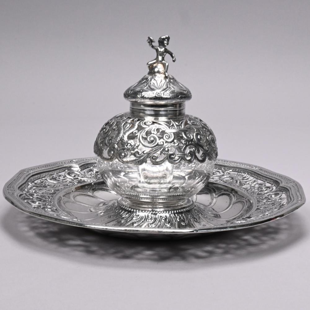 Silver and glass inkwell with an ornate domed lid and raised figurine finial, shown on a decorative round tray.