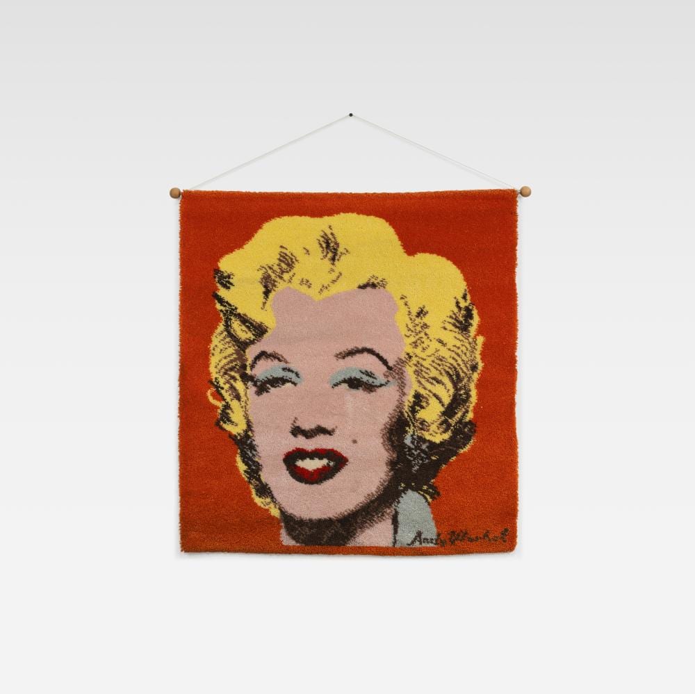 Square wall tapestry with a blonde woman’s face in pop-art style, hanging by four wooden corner pegs on a white background.