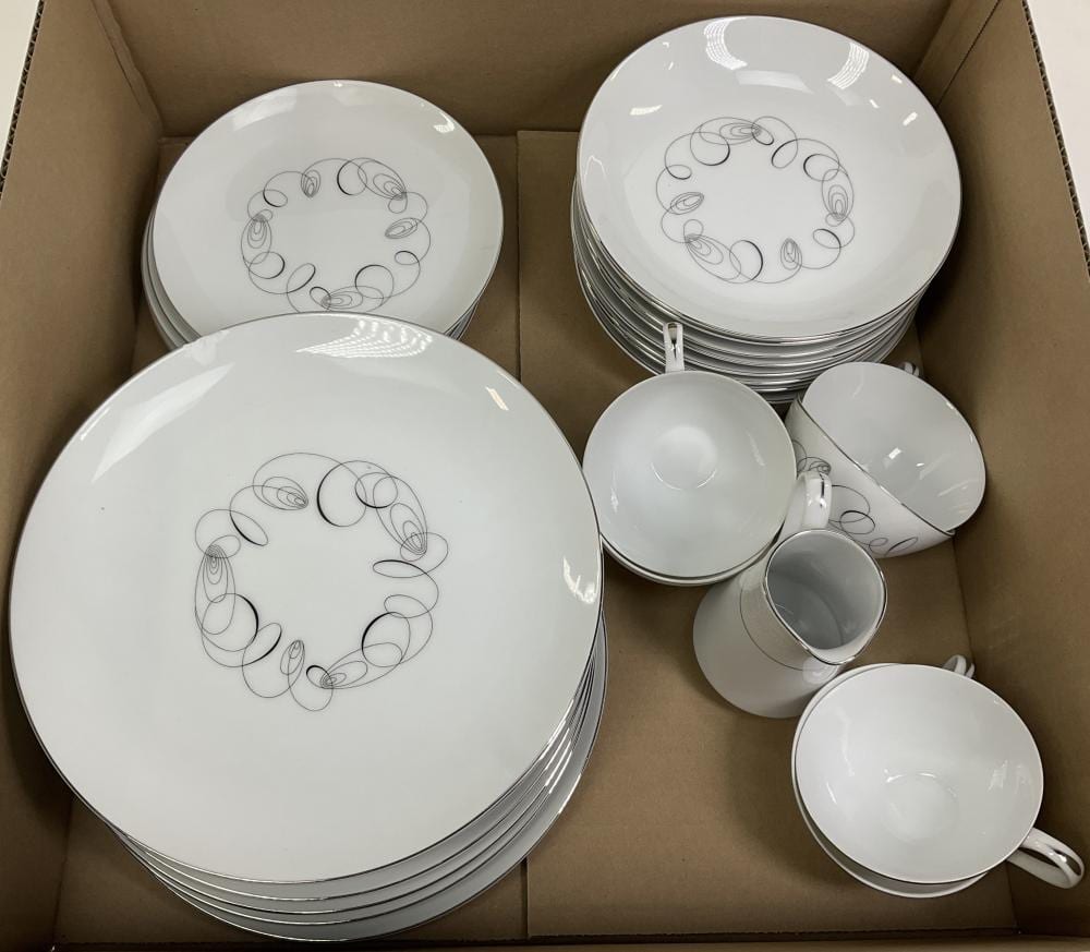 White porcelain plates and cups in a cardboard box with gray swirling line decorations around the center.