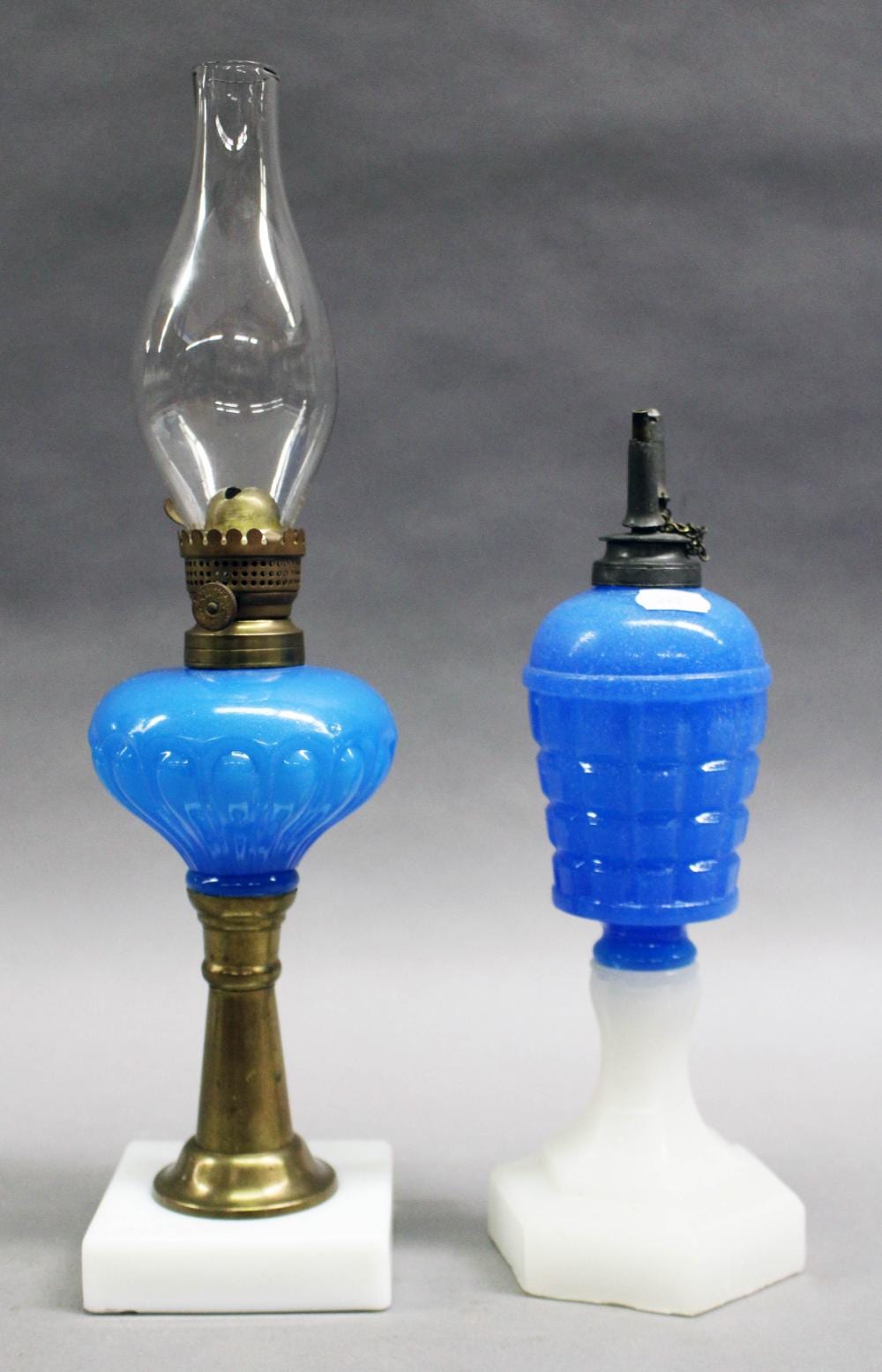 Two blue oil stand lamps with clear glass reservoirs and brass bases on white blocks against a gray background.