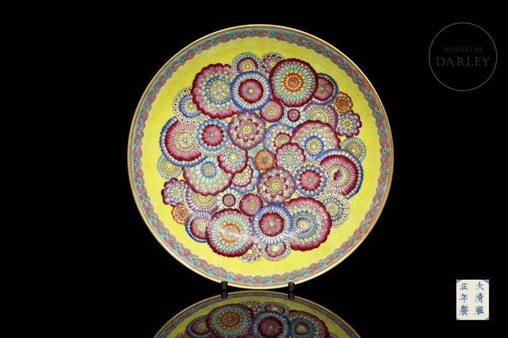 Glazed porcelain decorative plate with a yellow background and colorful circular floral motif in pink, blue, and purple, shown on a stand against a black screen