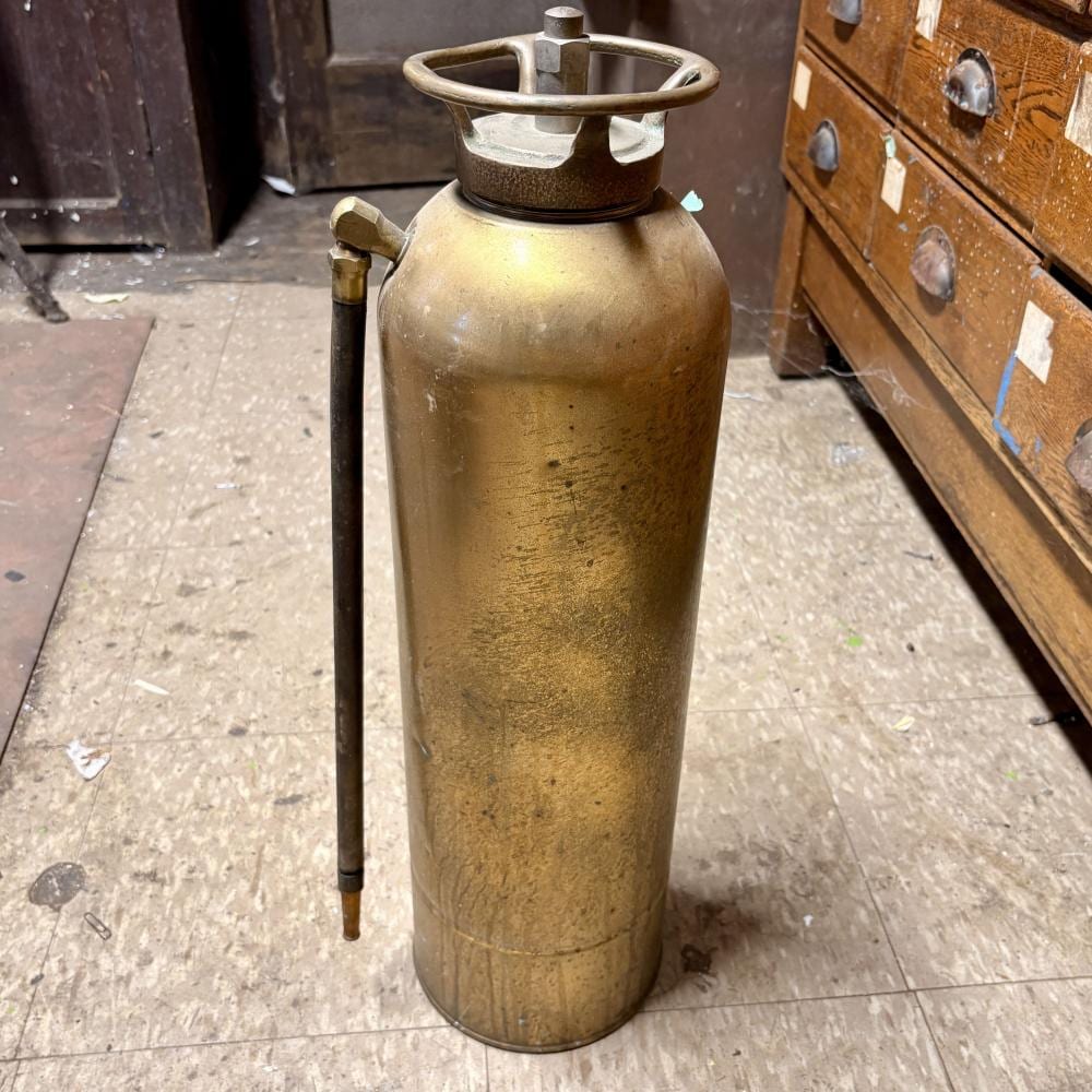 Antique-style brass fire extinguisher cylinder with an upright black handle and a circular metal valve guard on top.