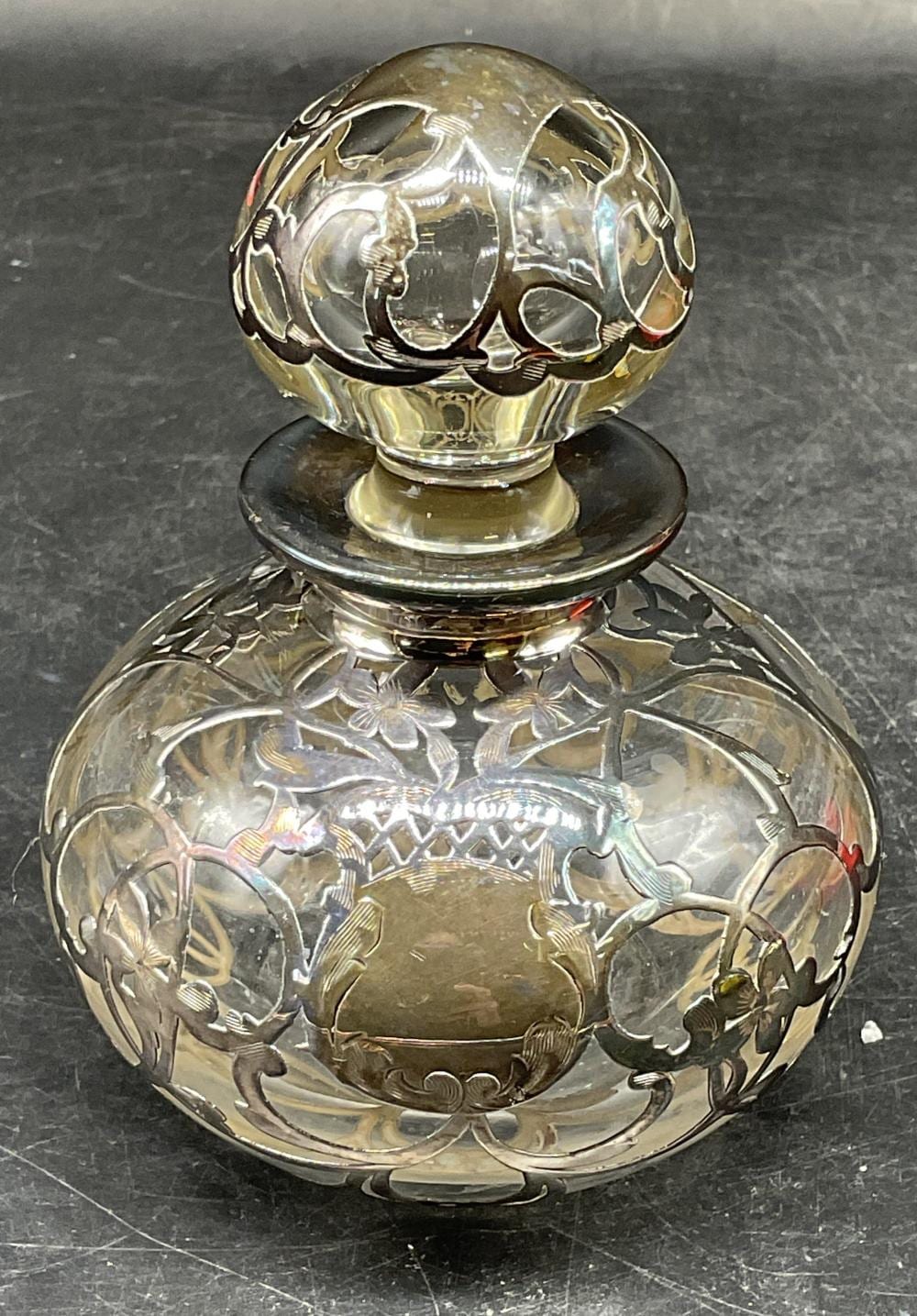 Close-up of an ornate glass perfume bottle with a silver-toned etched overlay, featuring floral and figural decorations.