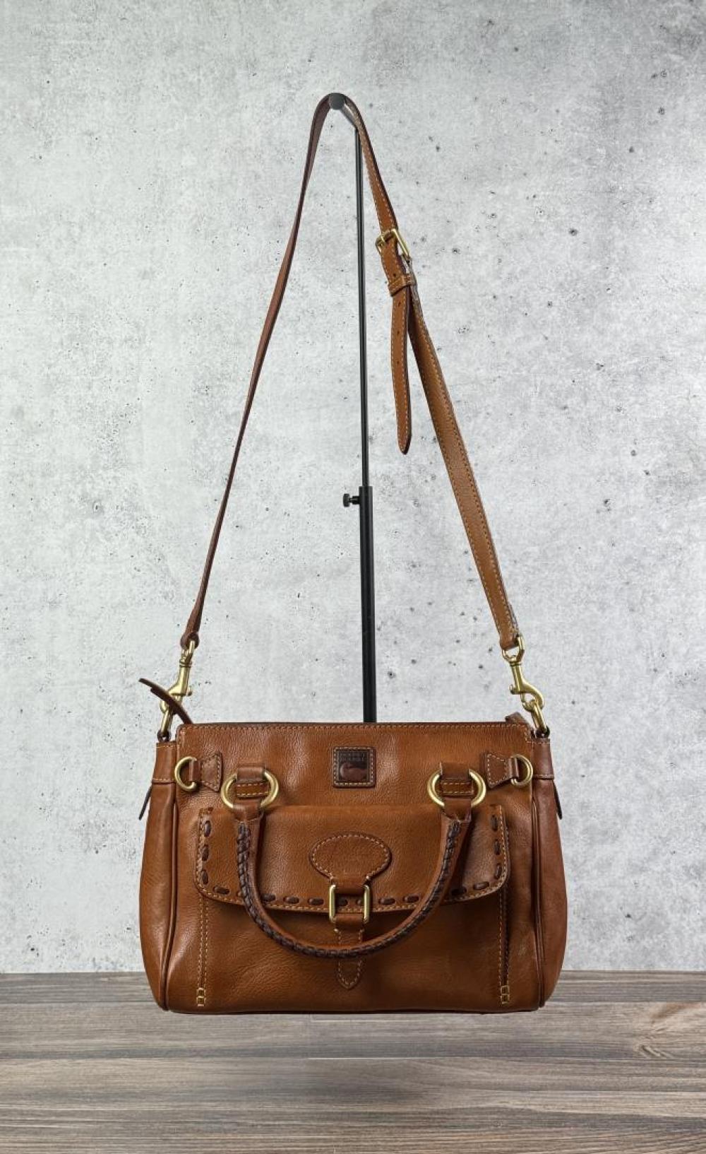 Brown leather handbag with gold-tone hardware, dual side straps, and a decorative front flap pocket shown on a stand.