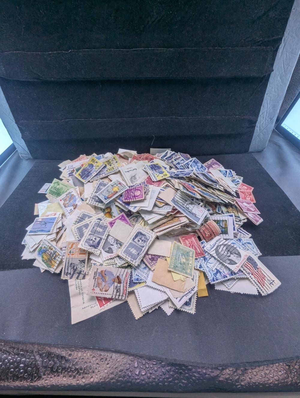 Pile of used United States postage stamps with colorful designs spread across a dark open box liner.
