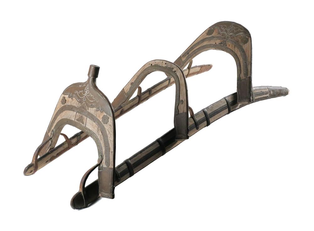 Antique camel saddle frame with decorative metal panels and arched handles, shown on a white background.