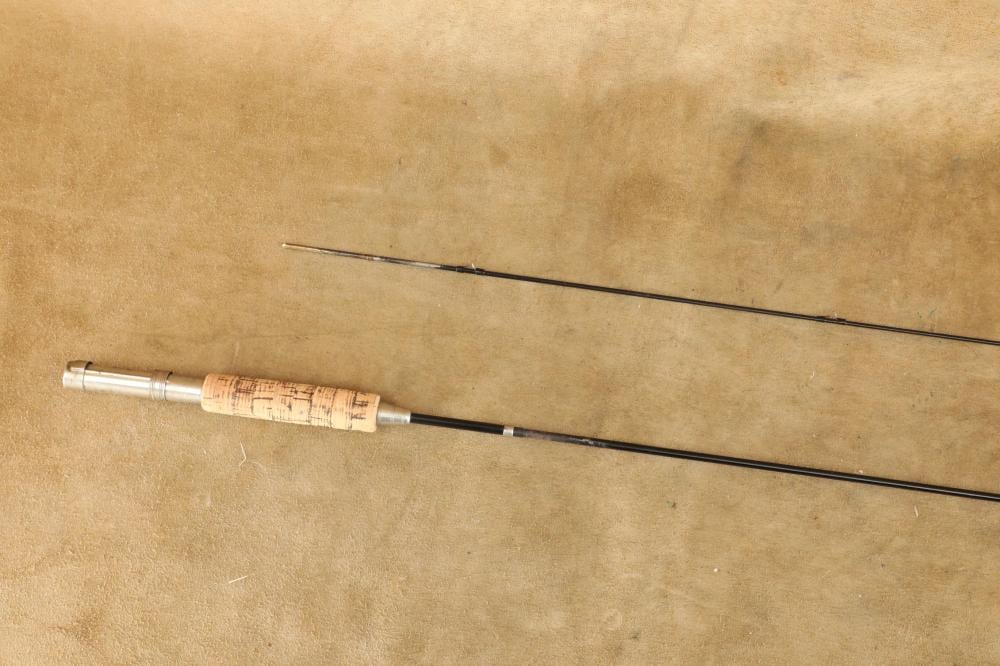 Two-piece fly fishing rod laid diagonally on a tan fabric background, showing the handle section and metal reel seat.