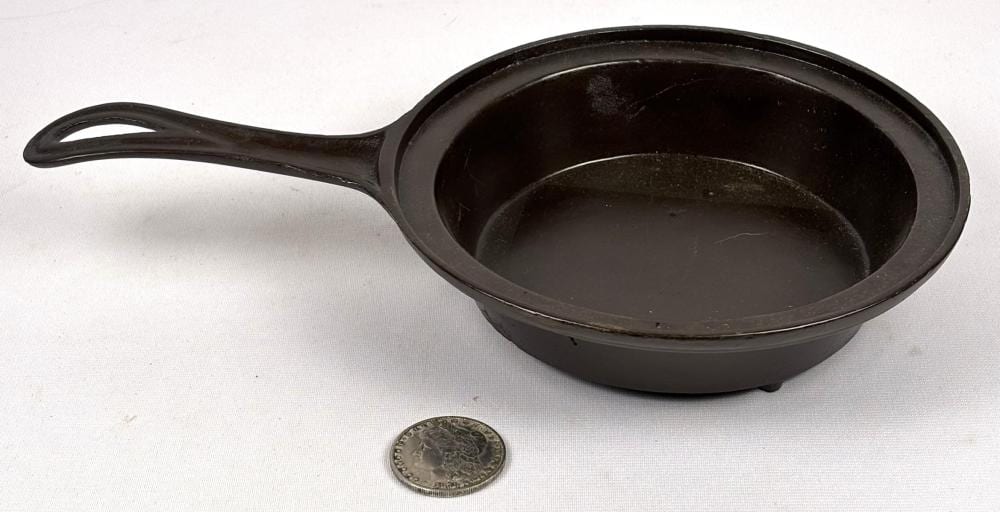 Black cast iron skillet with long curved handle, shown on a light background with a small coin for size comparison.
