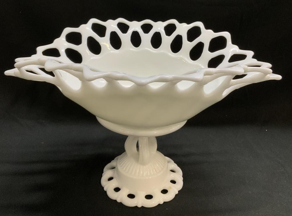White pedestal bowl made of milk glass with cutout lattice-like rim and looping handles on each side, on a black background.