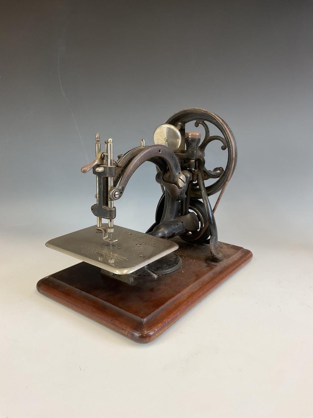 Antique metal sewing machine with a handwheel, needle mechanism, and metal presser plate on a wooden base.