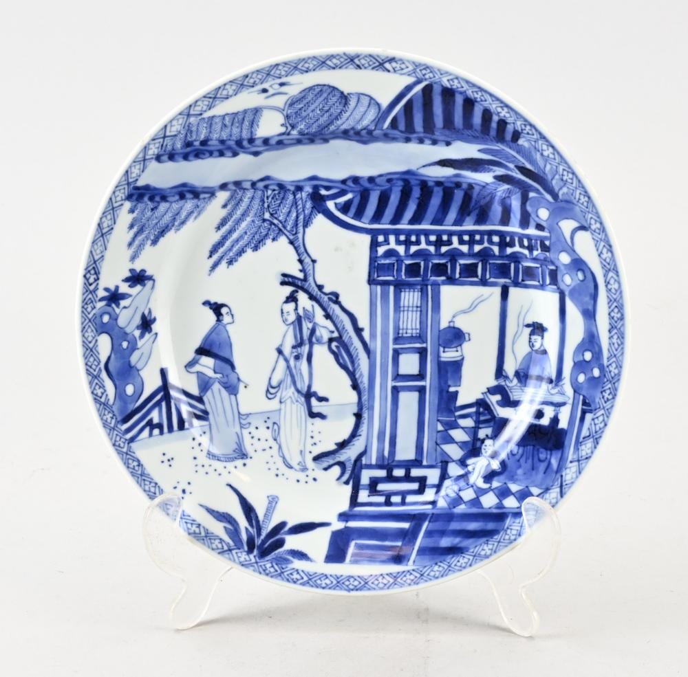Blue-and-white porcelain plate decorated with figures in a landscape and a building, displayed on a clear stand.