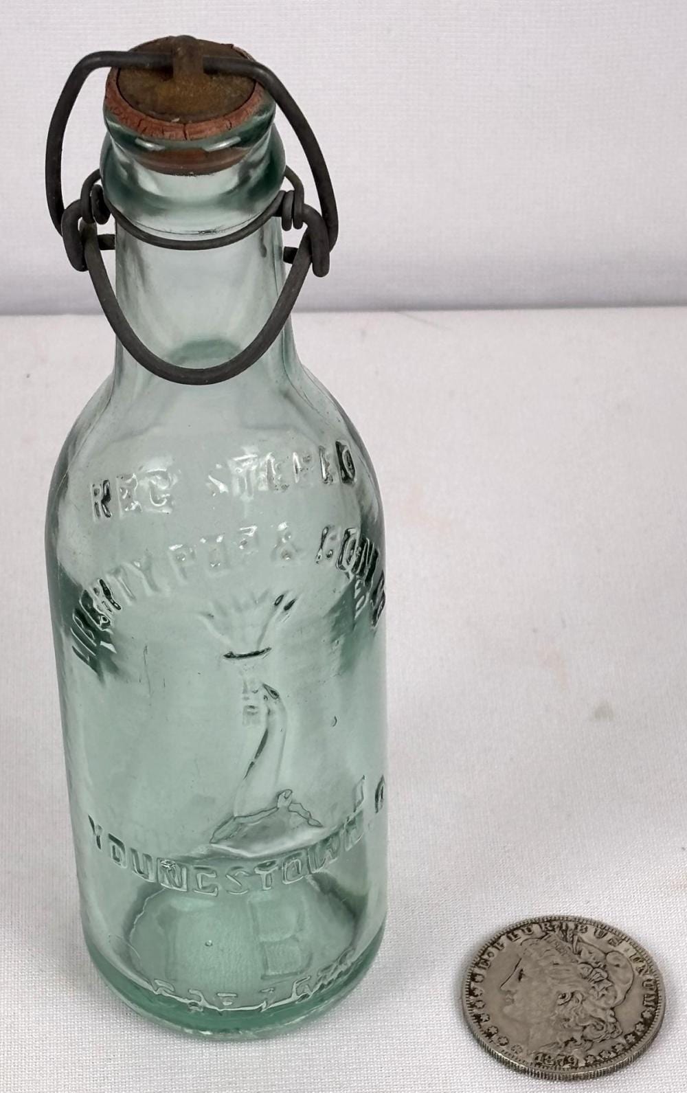 Green glass soda bottle with embossed lettering and a wire handle, shown next to a coin for scale.