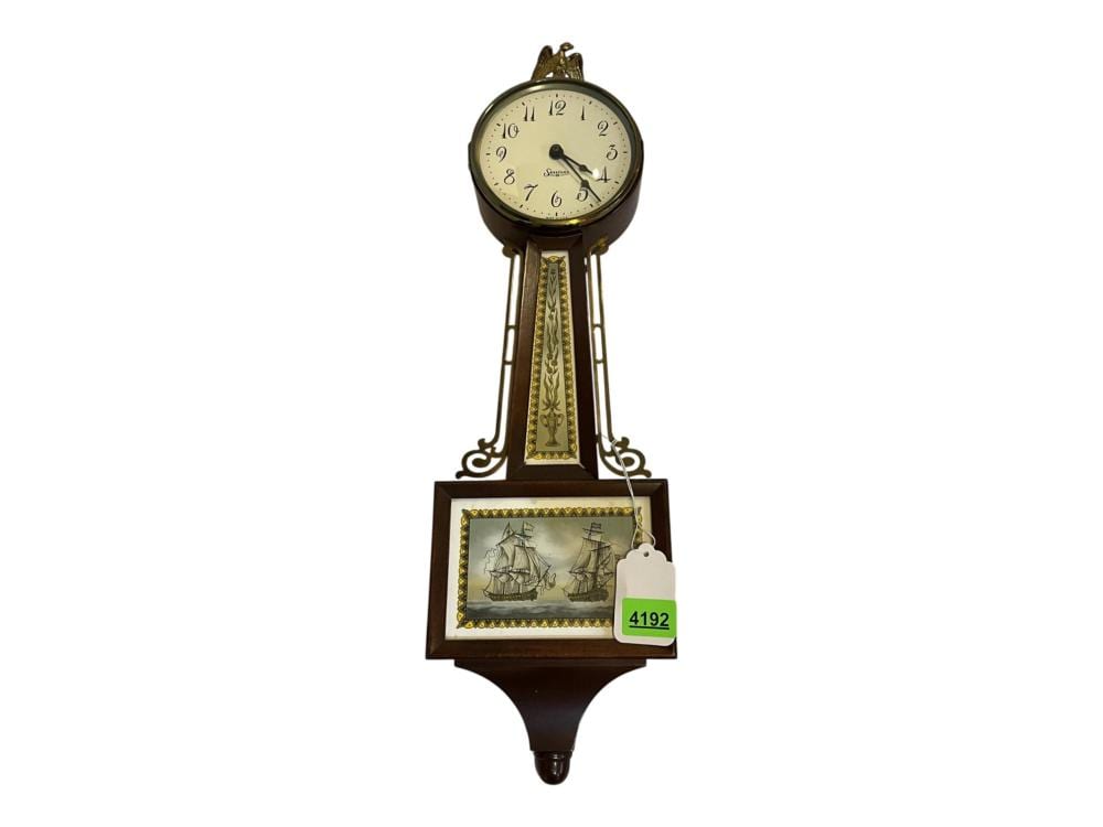 Wall clock with a round brass-toned face, black Roman numerals, pendulum case, and a small framed ship picture below the clock.