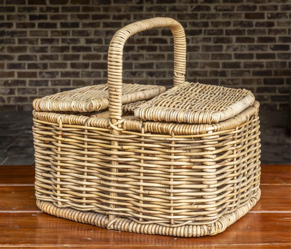 Vintage wicker picnic basket with a curved handle and woven lid-like top, sitting on a wooden floor in front of a brick wall.