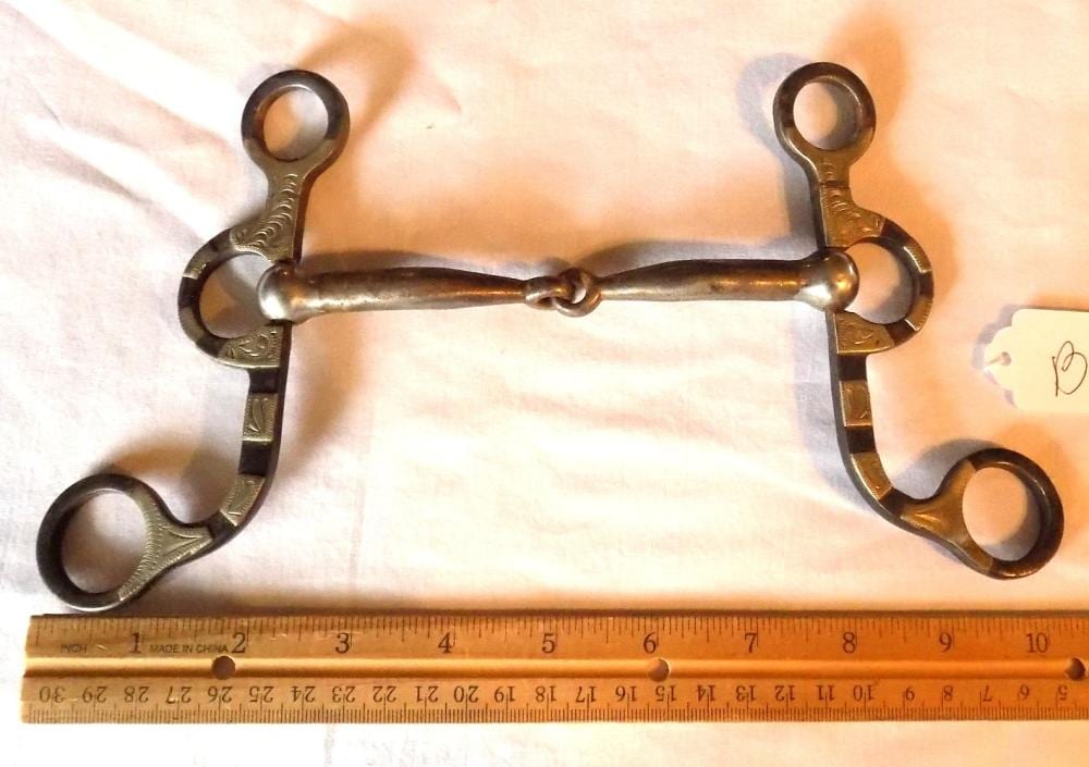 Brass horse riding bit with metal rings and engraved detailing, photographed next to a ruler for size.