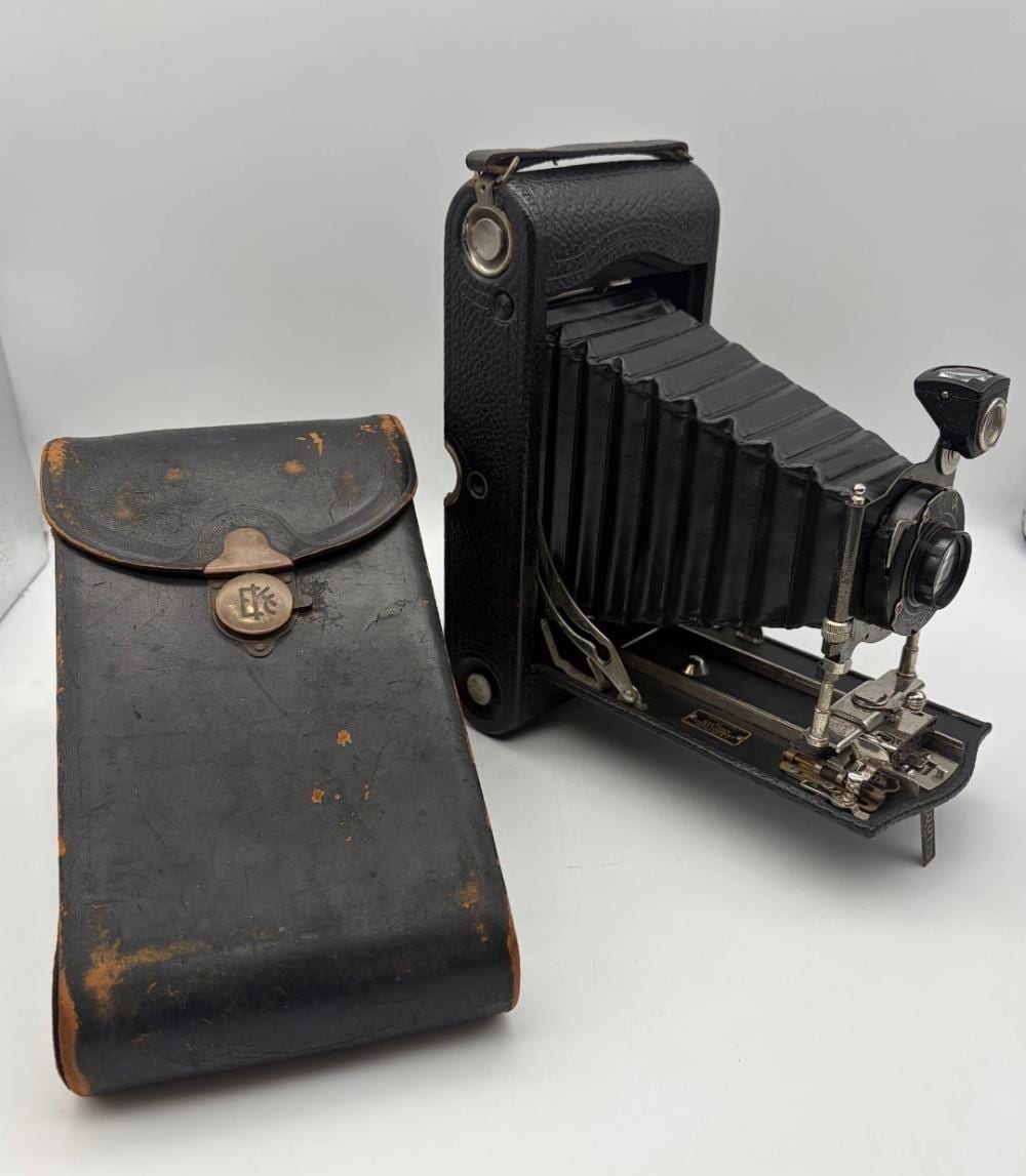Vintage folding camera with black leather body and bellows open, shown with a separate leather case on a white background.