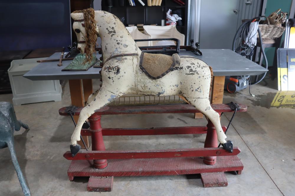 White wooden rocking horse with worn paint, curly mane, and saddle on a red rocking base in a workshop setting.