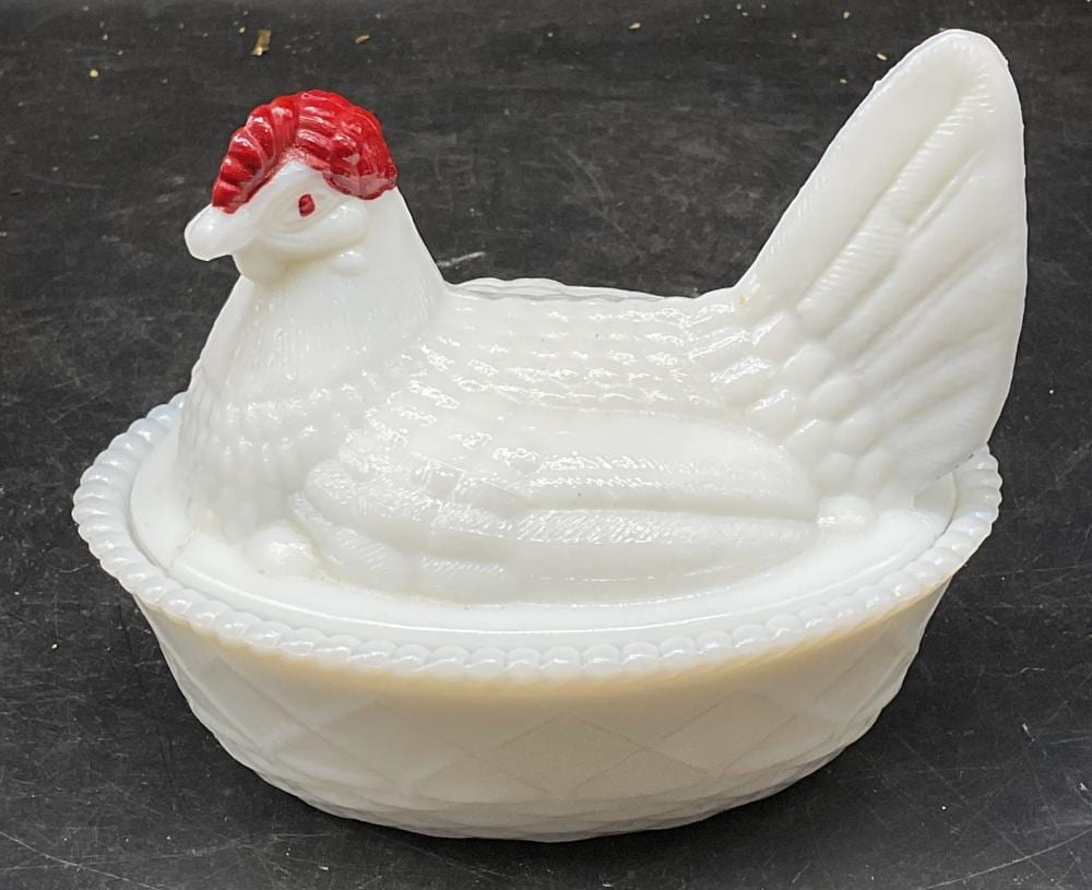 White milk glass hen-shaped covered dish with red comb and upright wing-like handle, shown on a dark tabletop.
