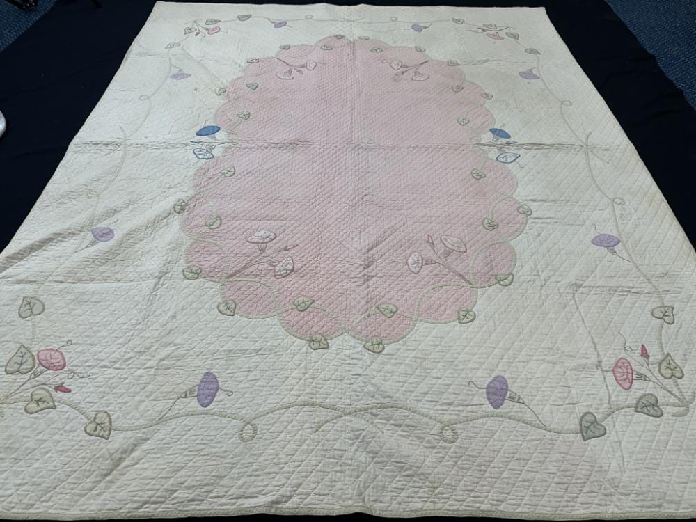 White quilt top with a central pink embroidered floral design and small pastel heart-like motifs around the border.