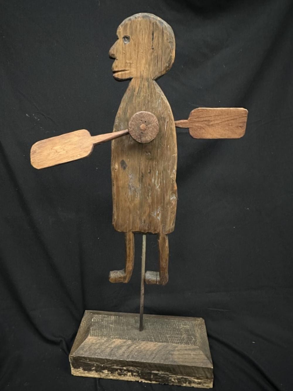 Carved wooden folk art figure on a base, shown in profile holding a wheel-like disc and rectangular arms over a dark background.