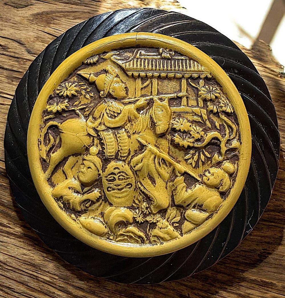 Close-up of a round two-color button with a gold-toned pictorial scene featuring an arched building, figures, and decorative floral motifs.