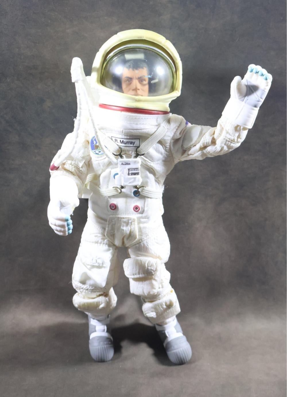 Vintage astronaut action figure in a white spacesuit with a helmet visor, arms raised in a greeting pose against a dark background.