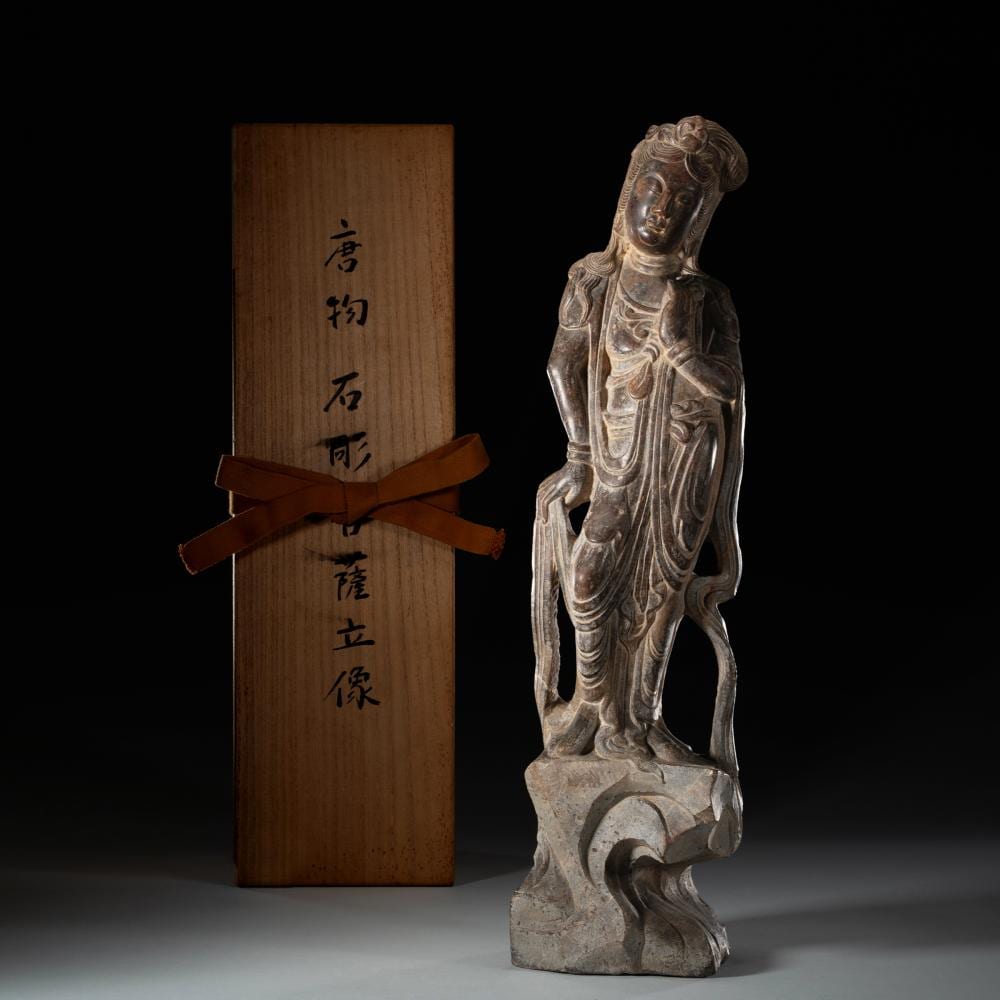 Silver-colored carved standing figure with flowing robes and a serene face, photographed beside a wooden inscription plaque tied with a ribbon.