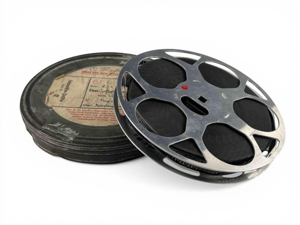 Spool of dark 16mm film with a metal film reel lying on top; label paper shows typed text and a red stamp.