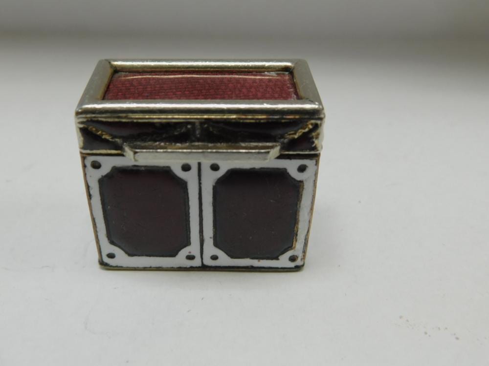 Small rectangular metal pill box with a hinged lid, guilloché-style paneling, maroon inner insert, and decorative corner and window-like motifs.