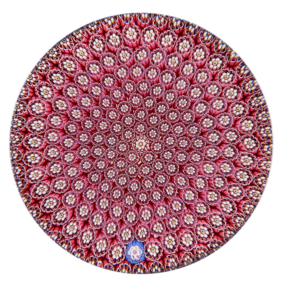 Round red millefiori glass paperweight with many small multicolored flower-like glass canes and a dark rim.