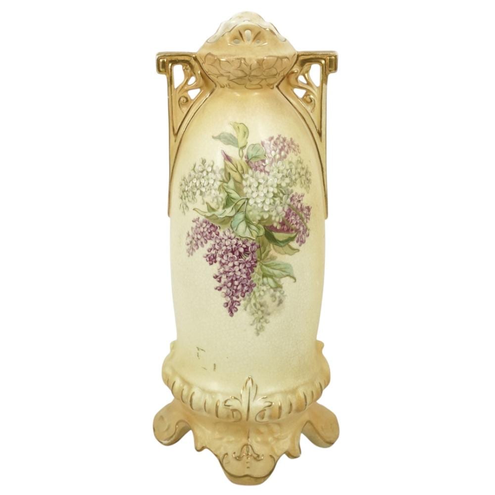 Porcelain vase with gilt handles and painted lilac and white flowers on a cream background.
