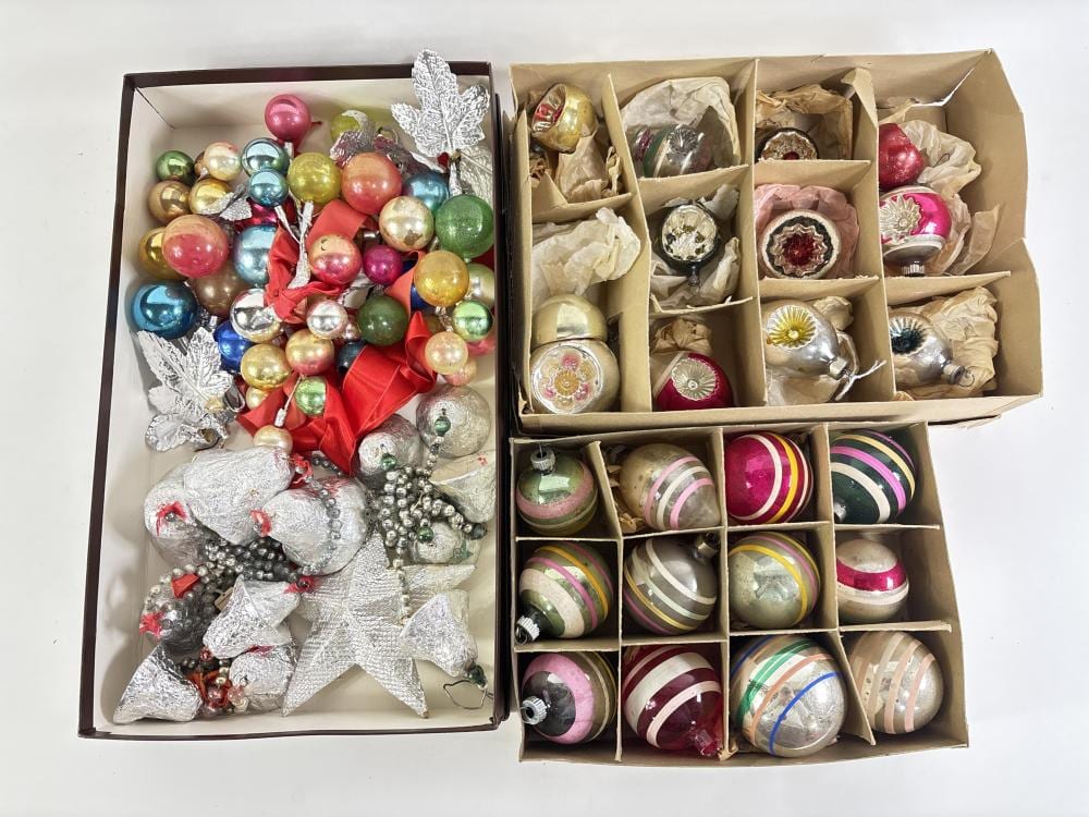 Two open cardboard boxes filled with vintage Christmas tree ornaments, including colorful ball ornaments and shiny metal star and leaf decorations.