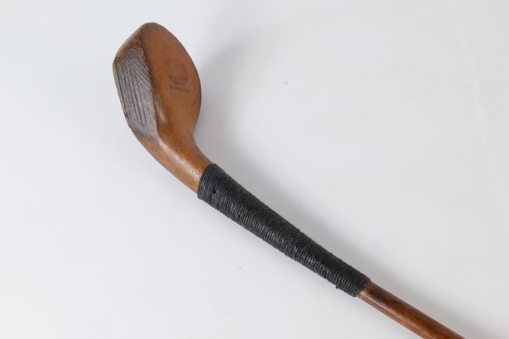 Antique wooden spoon-shaped golf club with a brown handle and a black grip, shown against a plain white background.