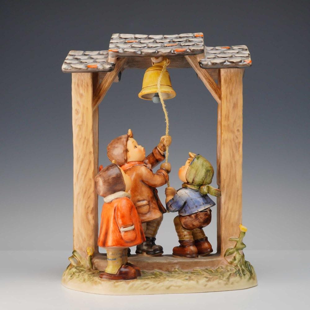 Ceramic figurine group under a wooden-style frame with a yellow bell, featuring three children holding and looking at the bell.