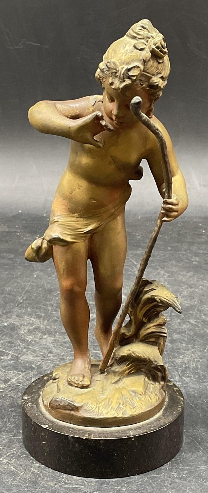 Gold-colored bronze-style sculpture of a nude female figure with a wreath and a dog or animal at her feet, standing on a round base.