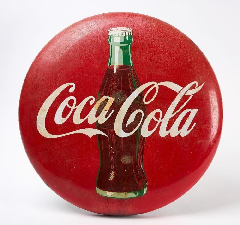 Round red porcelain button sign with a green Coca-Cola bottle graphic and white cursive “Coca Cola” text.