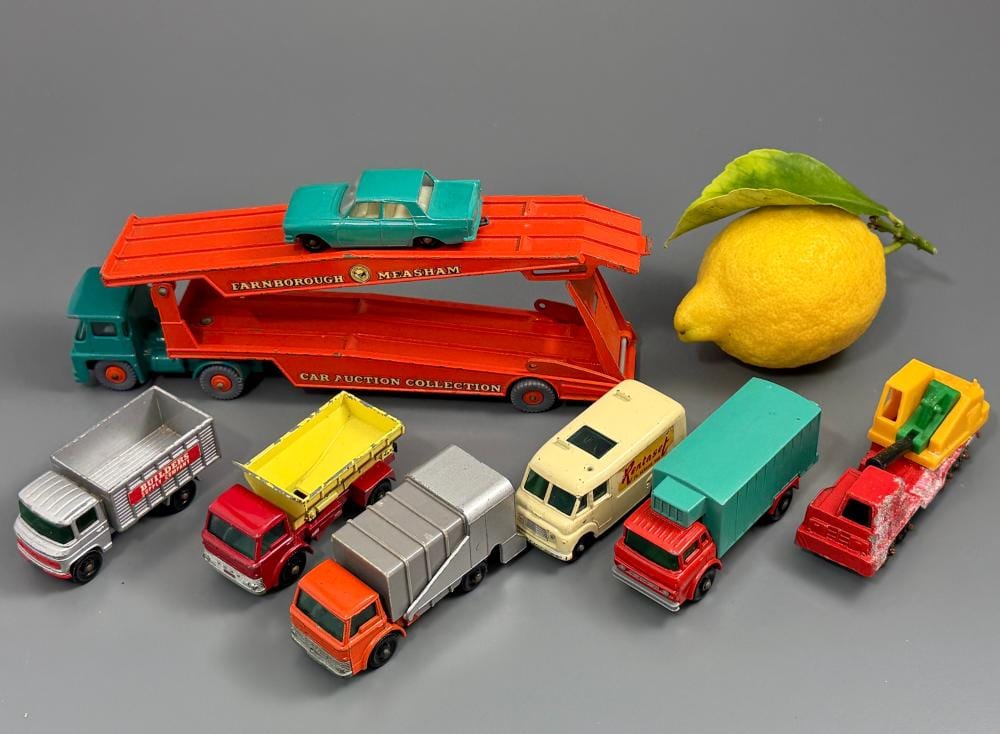 Assorted diecast toy car vehicles on a gray background, including a large red car transporter, smaller trucks, and toy car models; a lemon-shaped object sits to