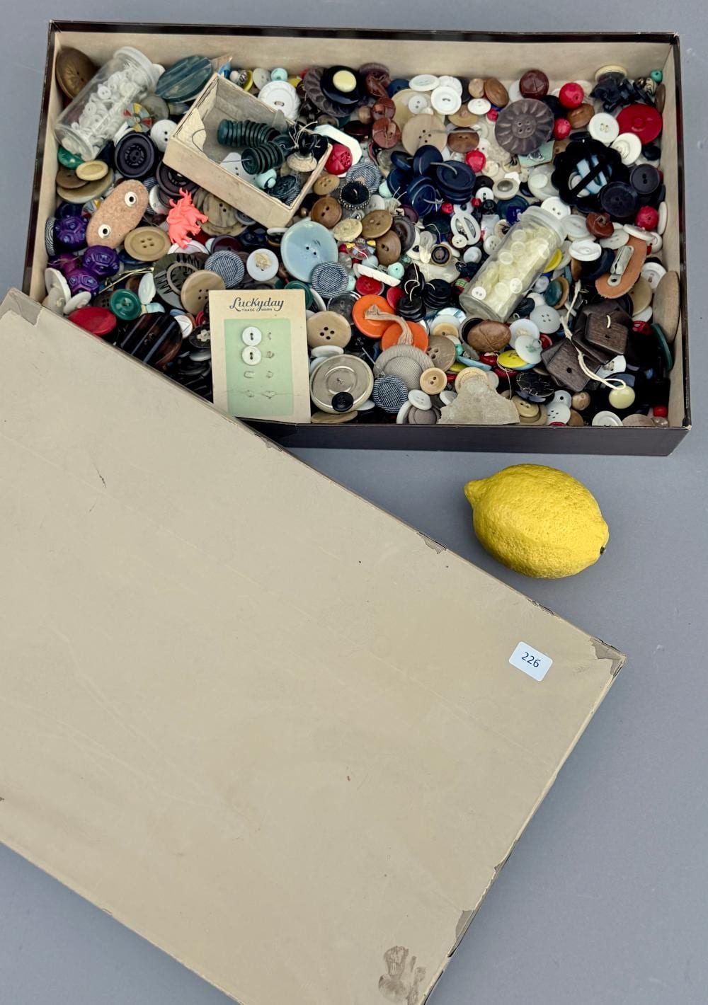 Open cardboard box filled with a mixed assortment of colorful sewing buttons and button pieces, including wood, plastic, and fabric-covered styles.