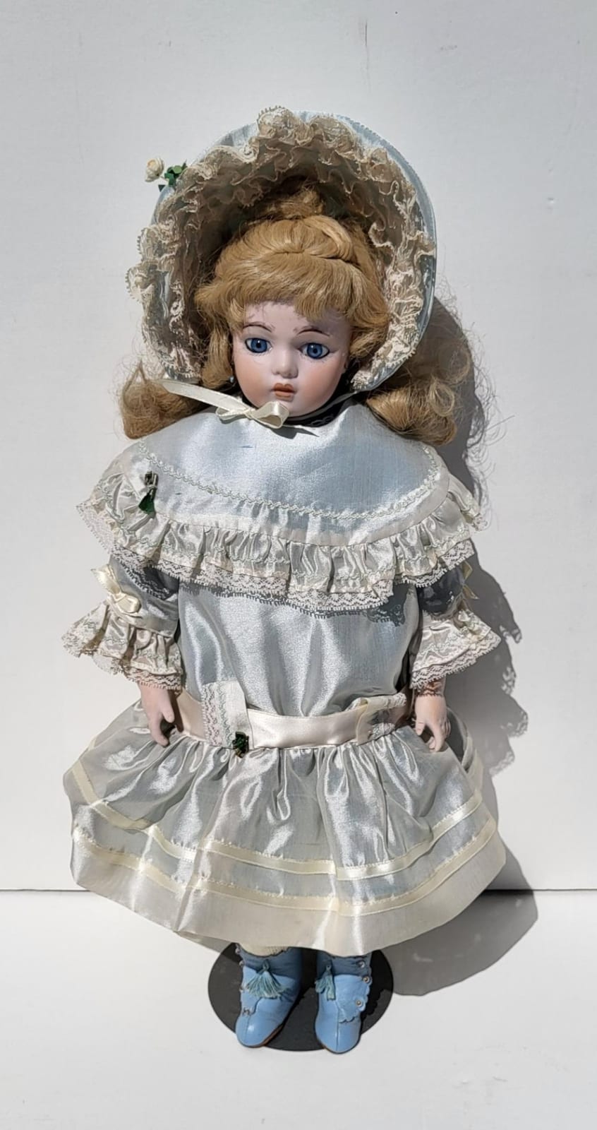 Porcelain doll with blonde hair, blue eyes, and a silver satin-style dress with lace trim and bonnet-like hat.