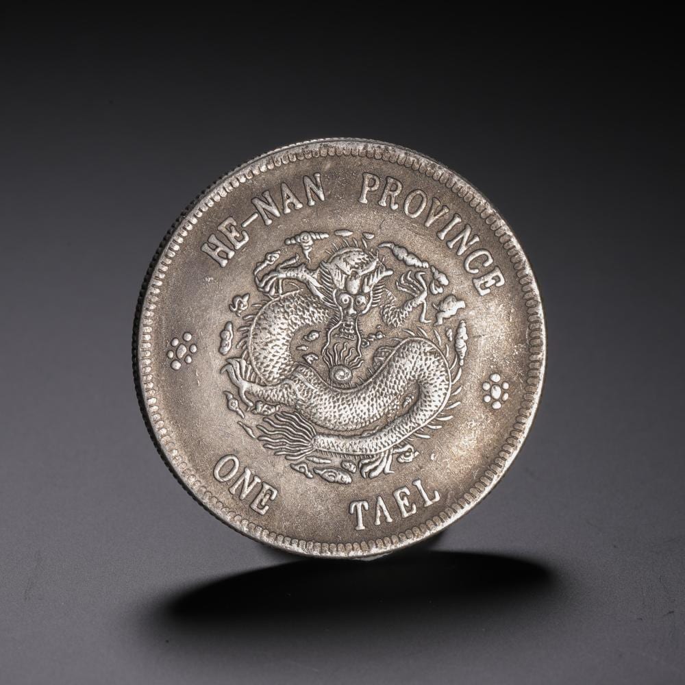 Close-up of a round coin showing a stylized dragon encircled by raised text, with “ONE TAEL” at the bottom.